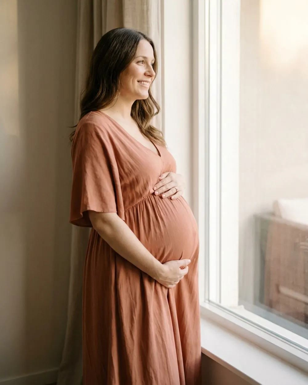 Une femme enceinte debout près d'une fenêtre, les mains sur son ventre